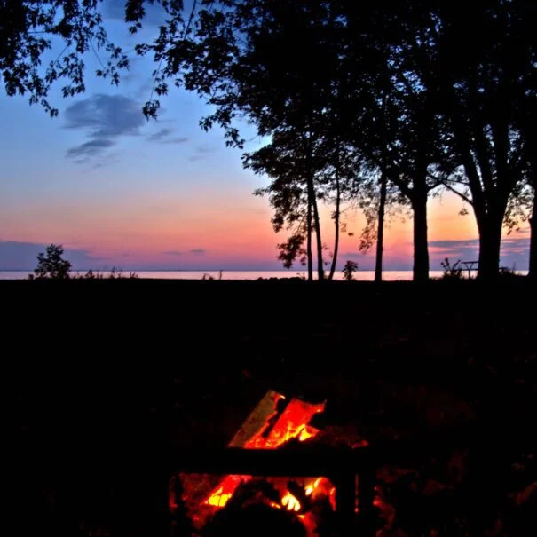 A campfire burning brightly at dusk, with silhouettes of trees and a colorful sunset in the background.