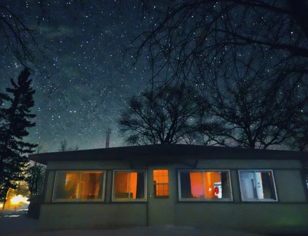 A cabin with lit windows, surrounded by trees and a starry night sky.