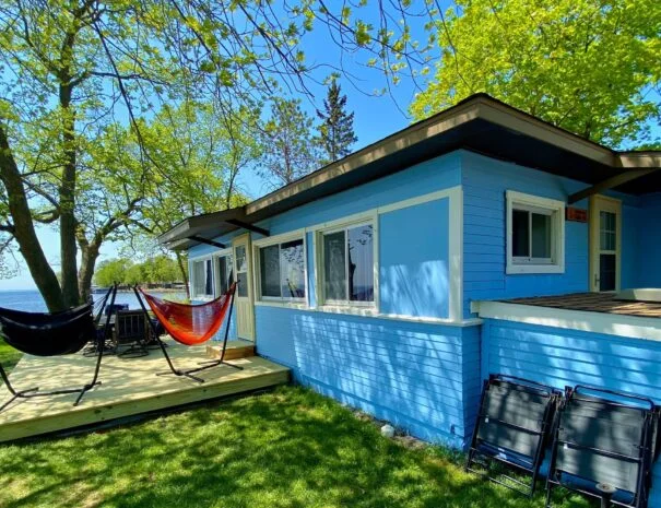 A blue cabin with a wooden deck and a hammock, surrounded by trees and a grassy lawn.